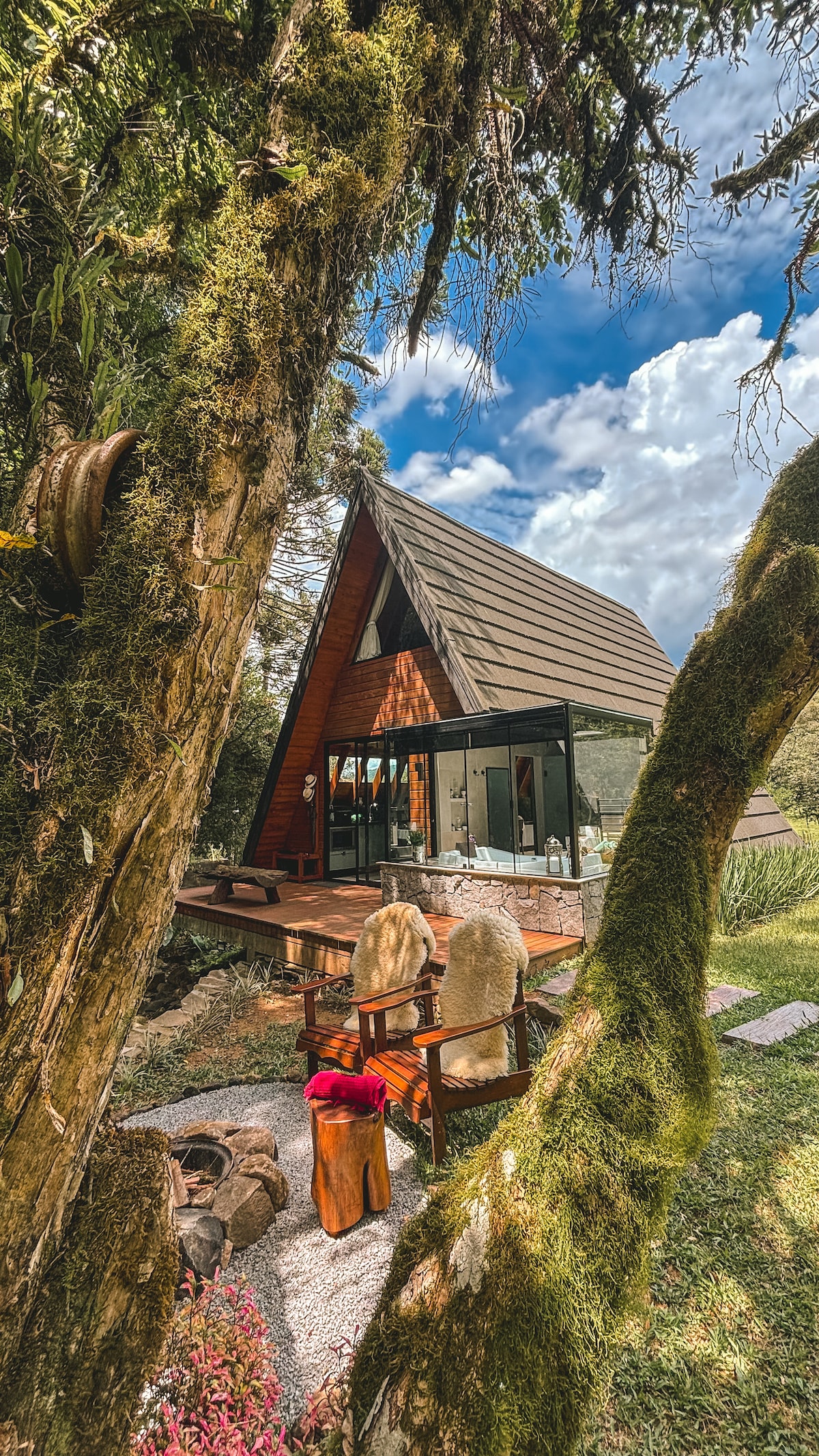 A modern cabin with a triangular roof is framed by lush greenery. Large glass windows reflect the surrounding landscape, inviting natural light indoors. A small outdoor seating area with rustic chairs rests on a pebbled path, enhancing the cabin's connection to nature.