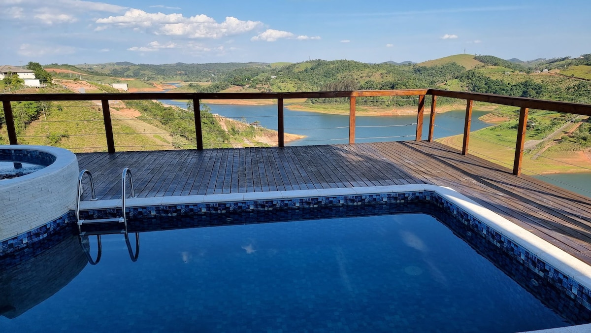 A spacious outdoor deck surrounds a private swimming pool, with a view of rolling hills and a serene lake in the background. The tranquil water reflects the blue sky, and a hot tub is positioned alongside. The railing offers unobstructed views of the natural landscape.