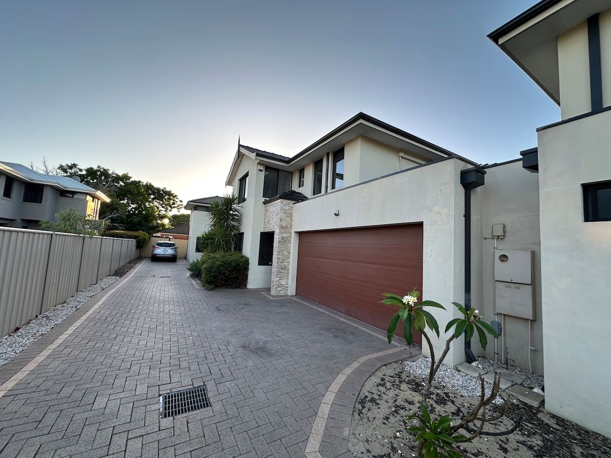 The exterior of a two-storey home is shown, featuring a driveway with pavers leading to a double garage with a brown door. Lush plants and gravel landscaping complement the façade, while neighboring homes are visible in the background under a clear sky.