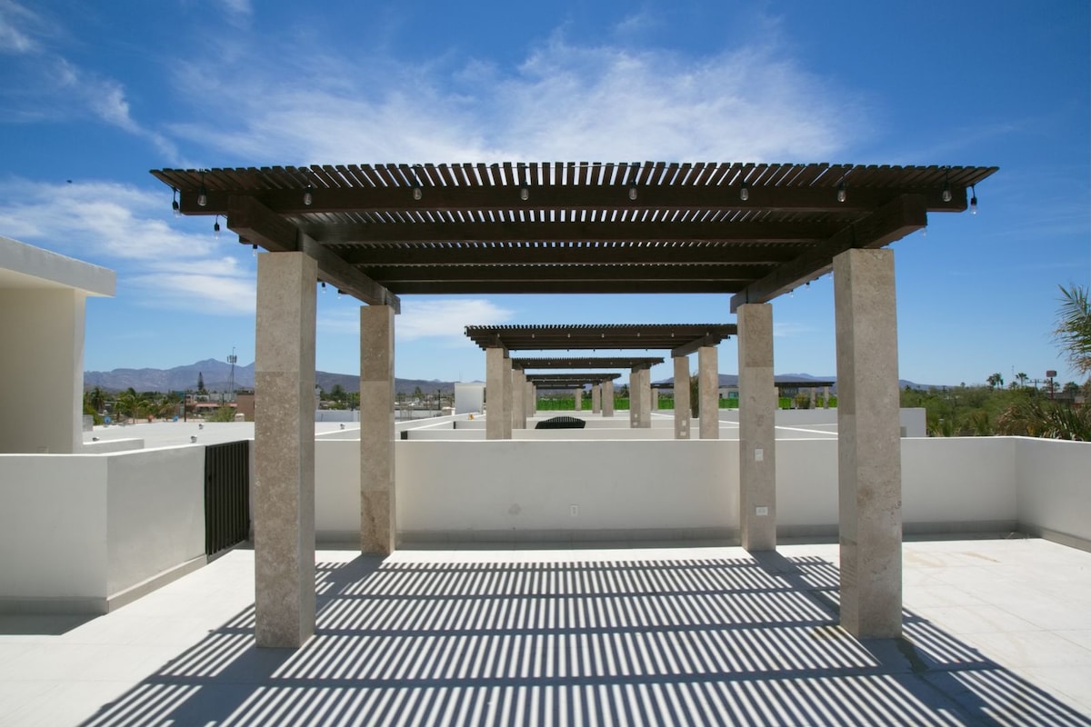 A spacious terrace is highlighted by a wooden pergola supported by stone columns. The area is bathed in natural light, casting patterned shadows on the ground. Clear skies and distant mountains create an open view, providing a sense of tranquility.