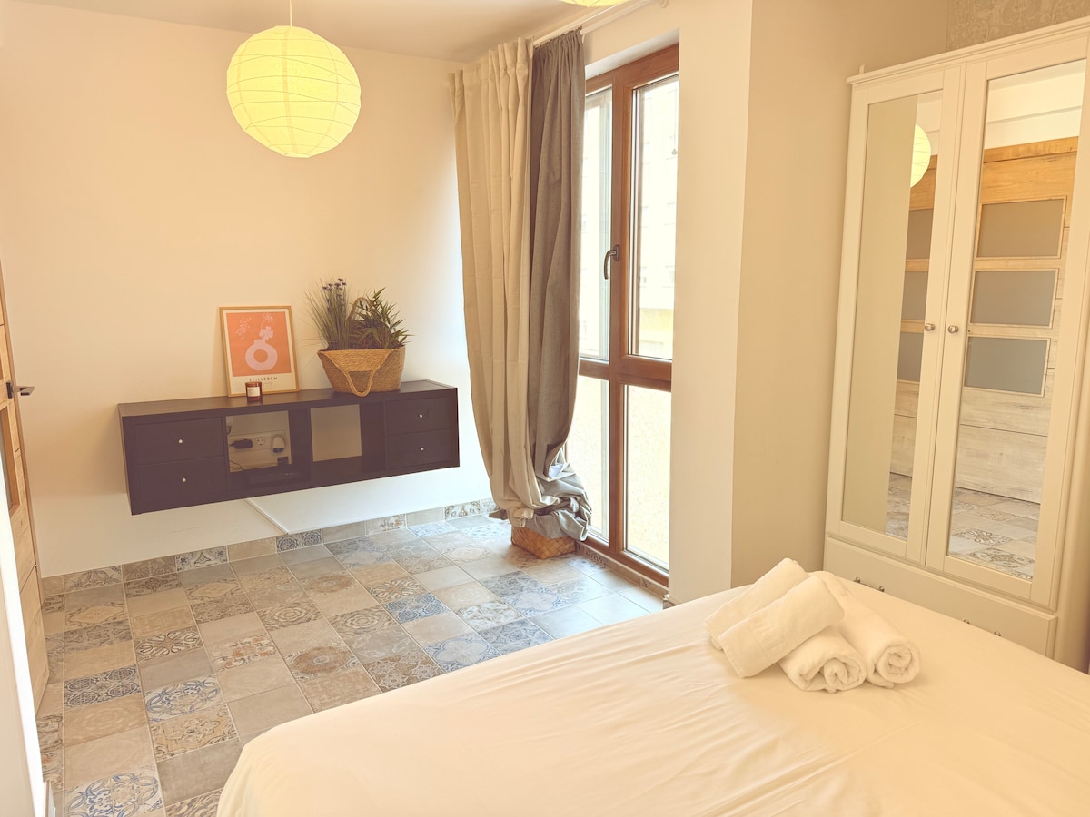 A well-lit bedroom features a spacious bed with neatly arranged towels. A modern wall-mounted cabinet is visible, along with a decorative basket. Large windows offer natural light and are complemented by soft curtains. The floor showcases patterned tiles, adding visual interest to the space.