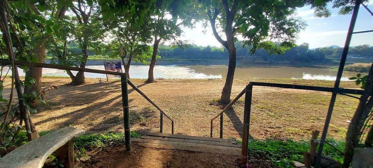 Steps lead down to a serene riverside area framed by leafy trees. The calm water reflects the surroundings, providing a peaceful atmosphere. An open grassy space is visible nearby, inviting relaxation or fishing activities.