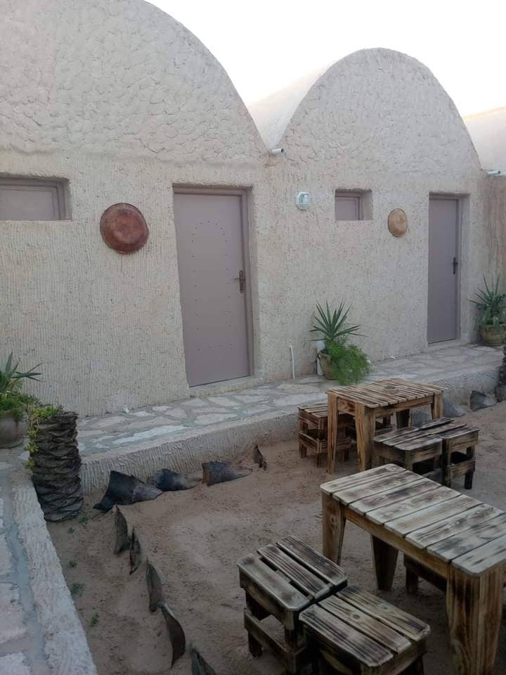 An exterior view of a unique structure featuring rounded, textured walls and two doors. Small wooden tables are arranged in a sandy area, surrounded by potted plants. The setting reflects a natural ambiance with earthy tones.