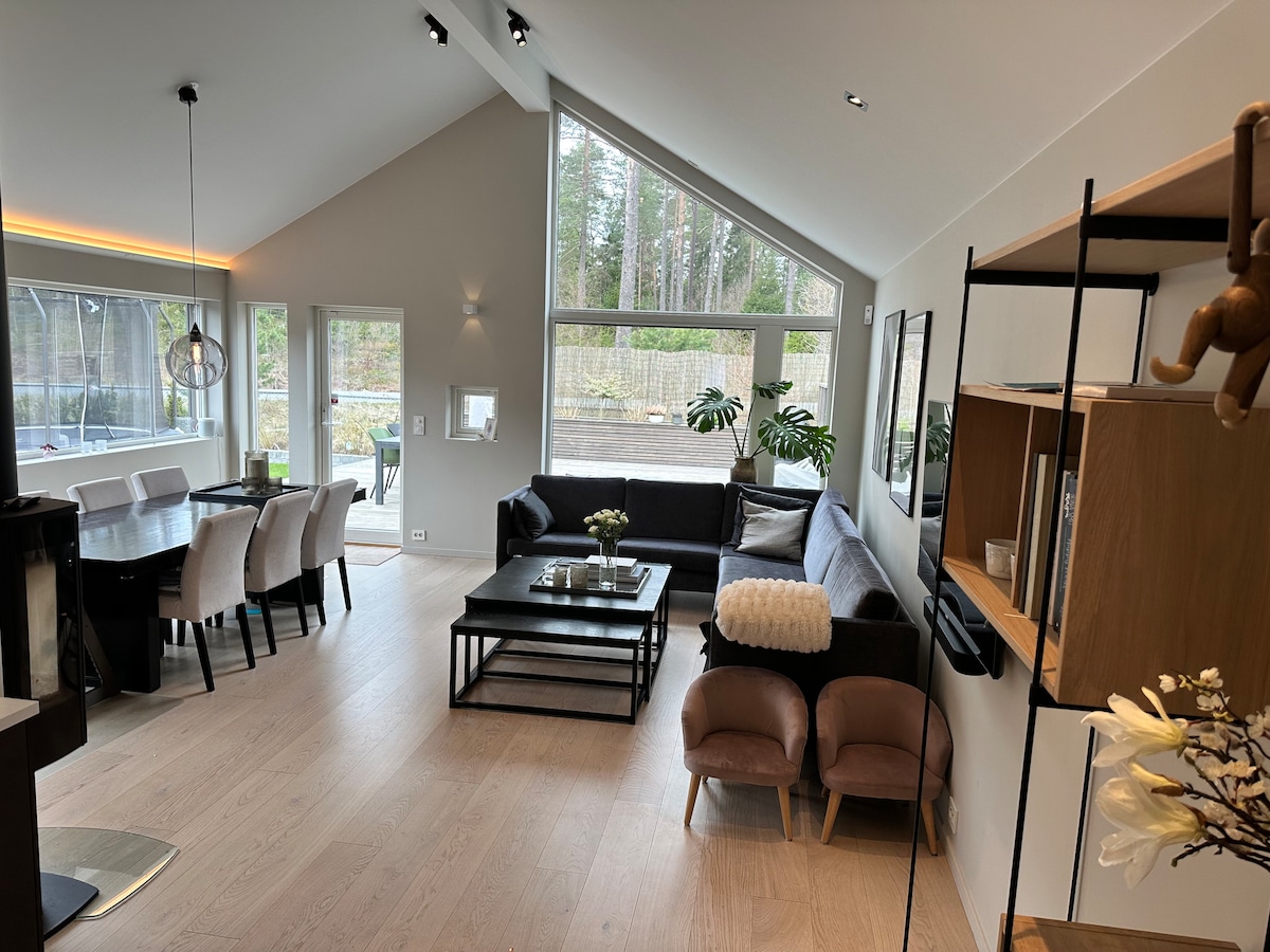 An open living area features large windows allowing natural light. A sectional sofa is positioned beside a coffee table, while a dining area with several tasteful chairs is visible. A combination of greenery and decor elements enhances the welcoming environment.