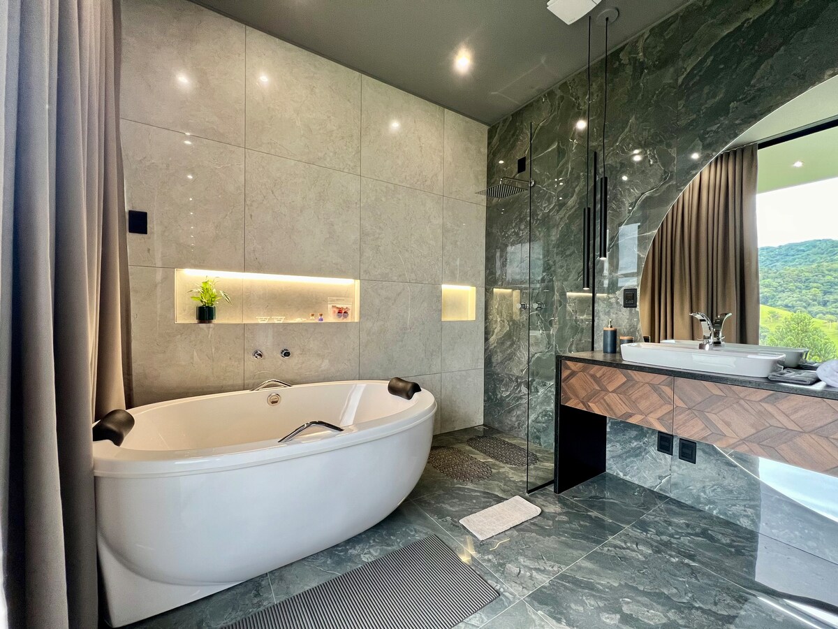 A modern bathroom features a spacious white soaking tub alongside a dual-sink vanity with a wood finish. Elegant green marble walls reflect natural light, while a large window offers a view of the surrounding nature. Soft lighting enhances the serene ambiance.