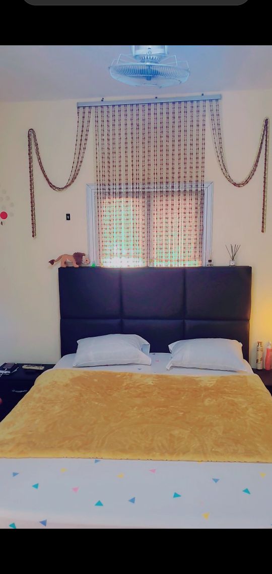 A cozy bedroom features a spacious bed covered in a yellow blanket. The headboard is dark, providing contrast against the light walls. A window dressed in patterned curtains allows natural light to enter, illuminating the room. Decorative elements include wall art and a small bedside lamp.