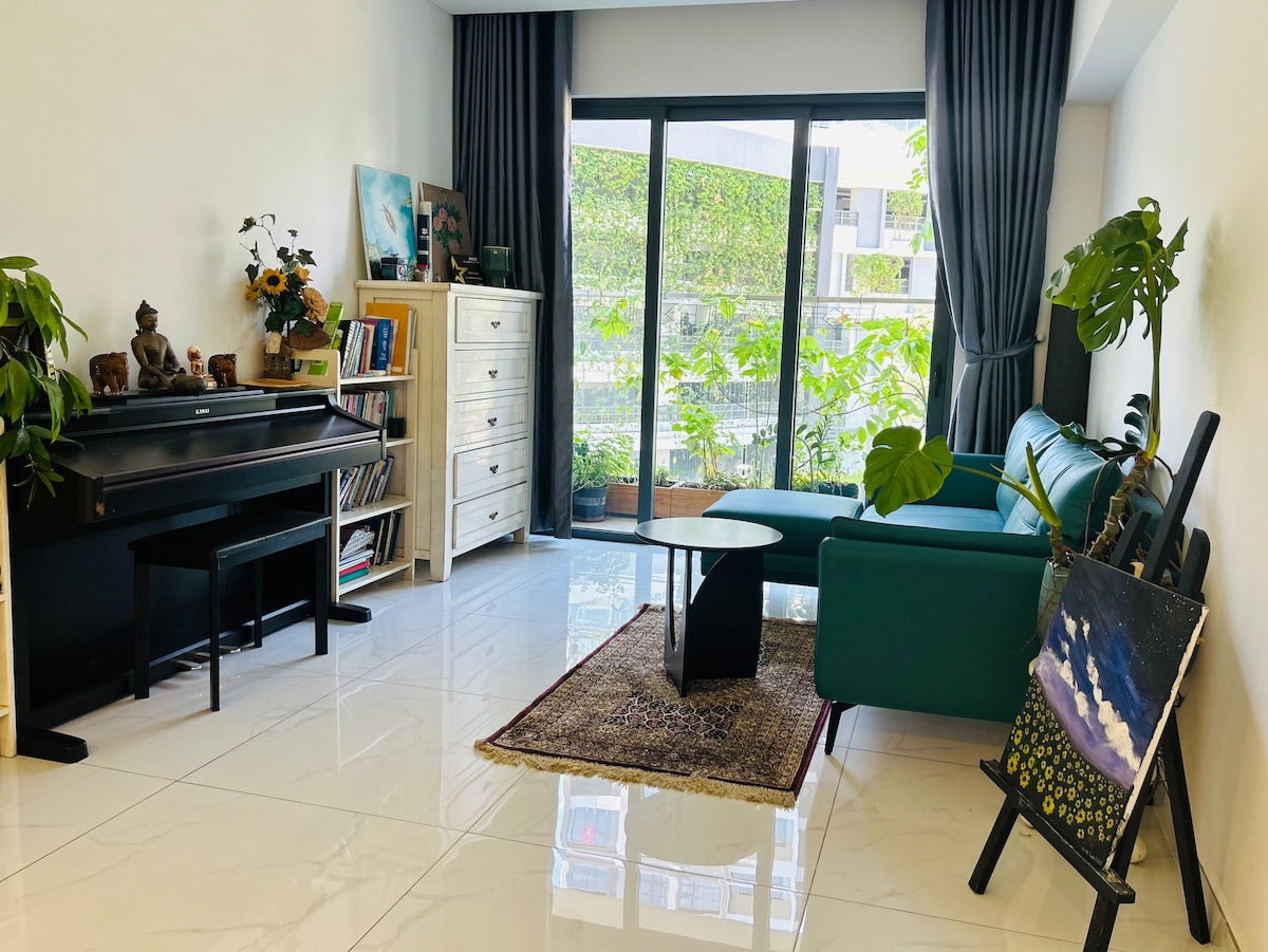 A modern living area features a black piano beside a white dresser filled with decorative items. A green sofa faces a sleek black coffee table, complemented by a woven area rug. Large glass doors provide a view of an outdoor green space, enhanced by natural light.