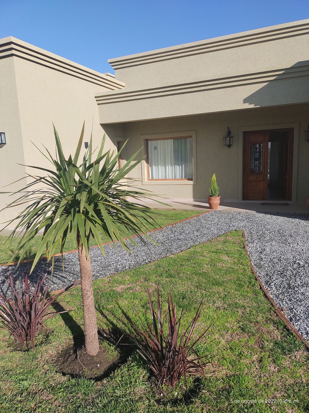 The entrance of the house is framed by a well-maintained path lined with gravel and lush greenery. A small palm tree and ornamental plants are positioned strategically, leading to a wooden door adorned with decorative accents. Natural light enters through a large window nearby.