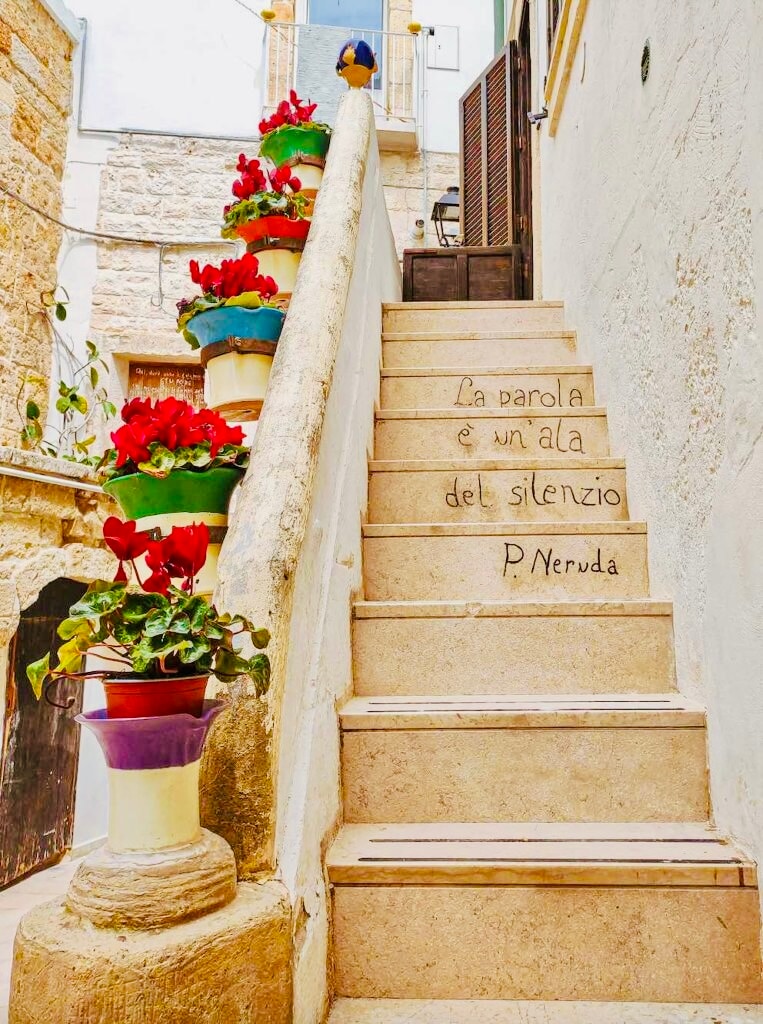A stone stairway leads upward, embellished with vibrant flower pots housing blooming red plants. An inscription on the steps reads, 'La parola è un'ala del silenzio P. Neruda,' integrating art into the natural surroundings.