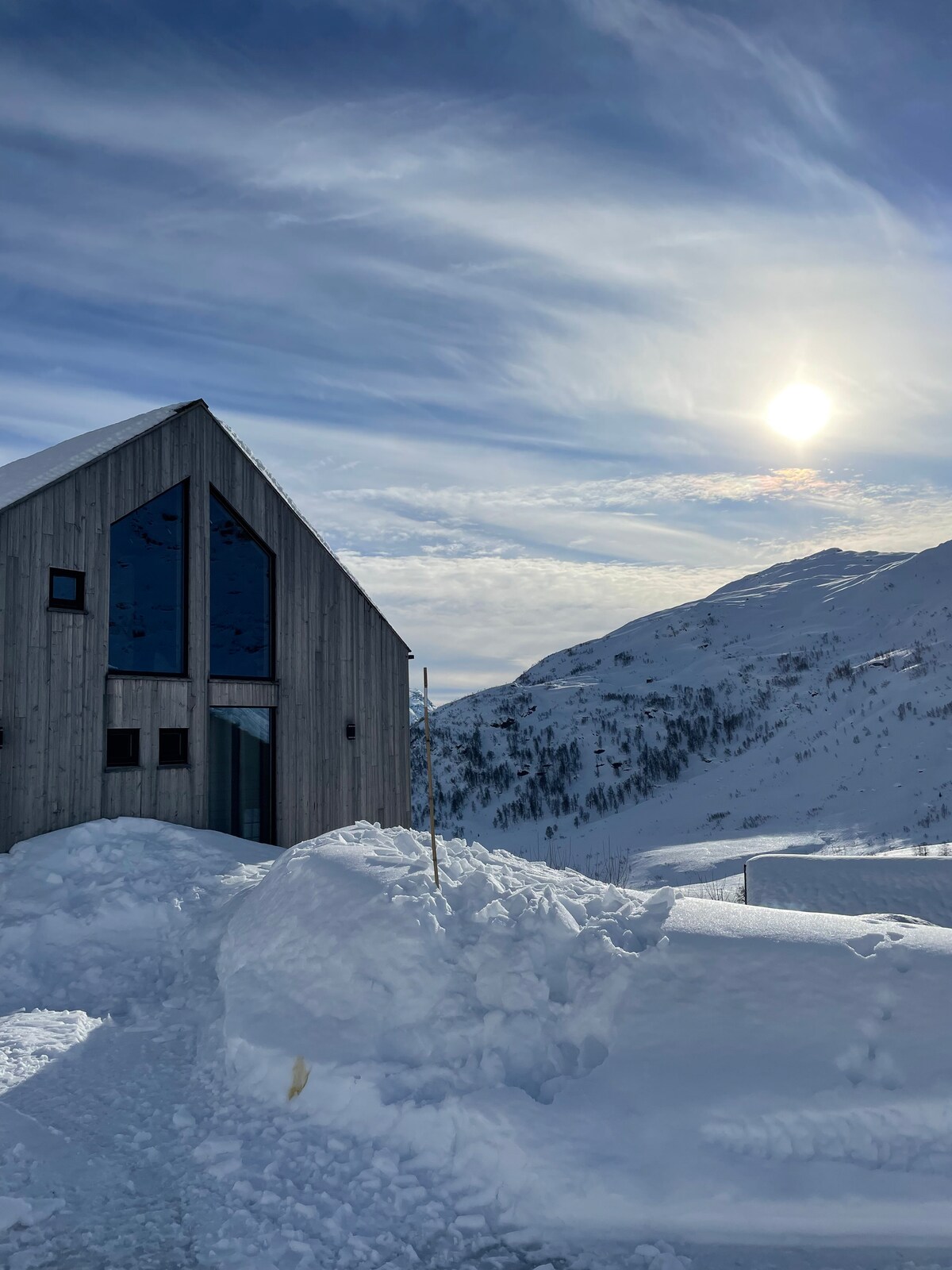 A unique mountain cabin is set against a backdrop of snow-covered hills and a bright sky. Large reflective windows create visual interest, while the sun is seen rising or setting on the horizon. Snow drifts frame the cabin, highlighting the winter landscape.