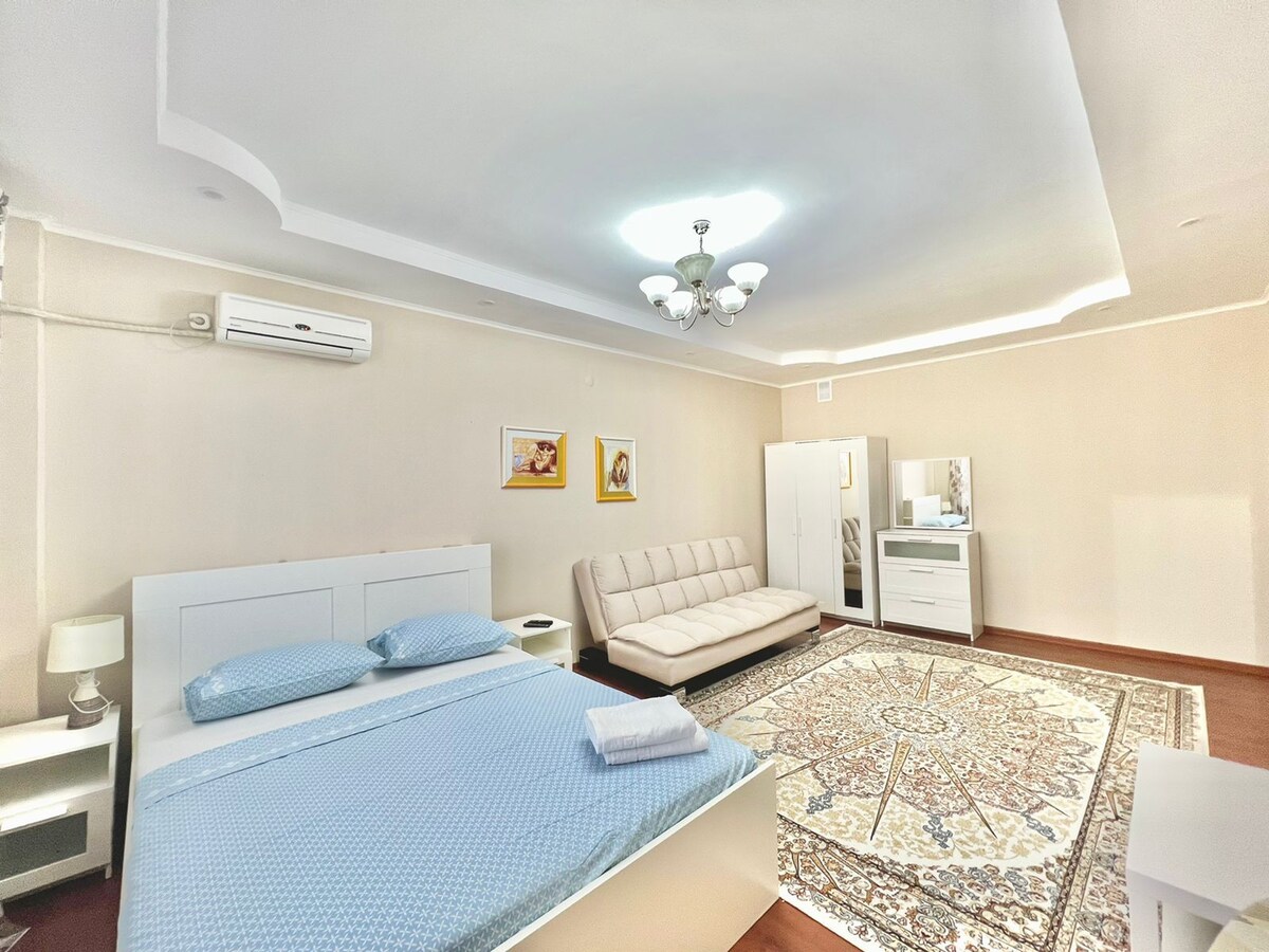 A spacious bedroom is presented, featuring a large bed with a light blue blanket and matching pillows. A soft cream sofa sits adjacent, while a white dresser and wardrobe are positioned nearby. Warm lighting from a chandelier complements the neutral wall tones and decorative area rug.