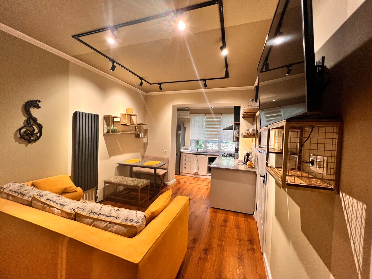 A spacious living area features a mustard-yellow couch positioned near a wooden dining table. Open shelves display decorative items, while a modern kitchen is visible in the background, equipped with a countertop and cabinetry. Soft lighting enhances the inviting atmosphere.