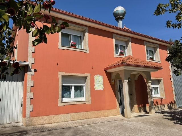 The exterior of the house is featured in warm orange tones, accented by stone detailing. Bright flowers are visible in window boxes. A canopy above the entrance provides shelter, while a water tower stands above the roof, completing the welcoming facade.