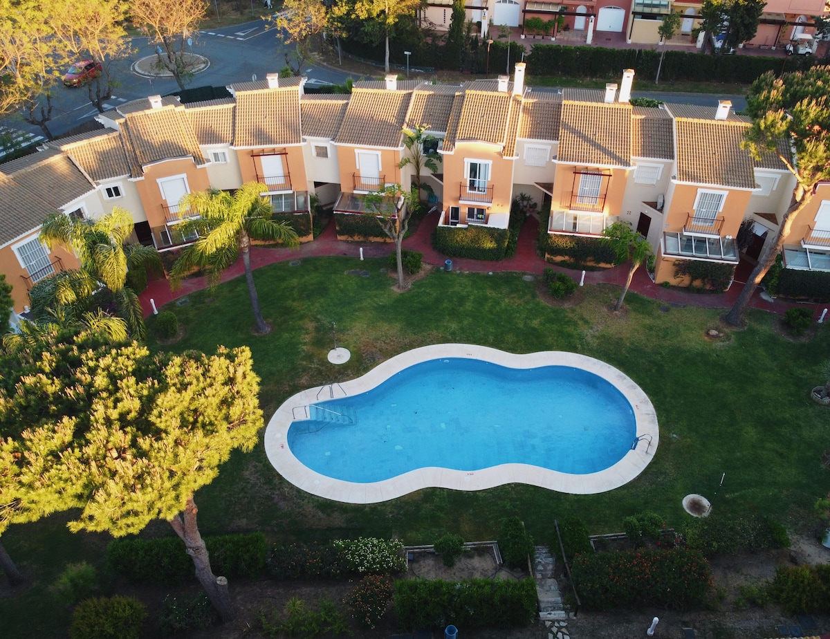 An aerial view of a cluster of townhouses surrounding a kidney-shaped swimming pool. Lush greenery and palm trees are visible in the garden area, providing a natural frame for the pool, which features a set of stairs leading into the water.