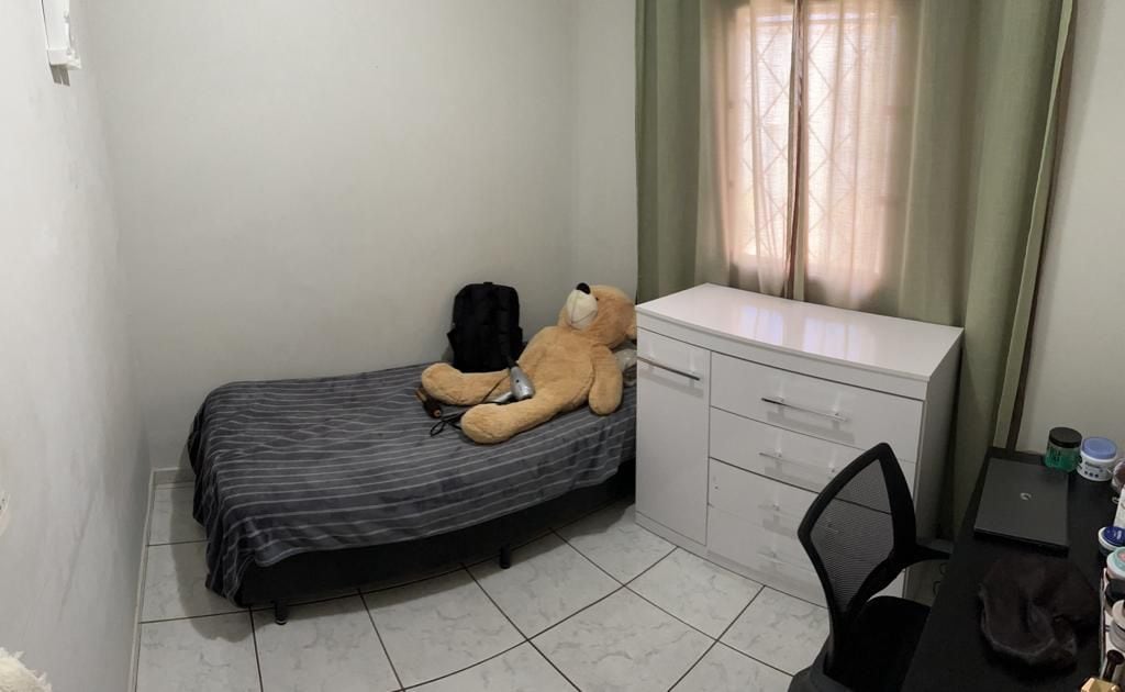 A simple sleeping area features a twin bed with a gray striped blanket and a large teddy bear resting on it. A chest of drawers in white sits nearby, while a chair and desk with various containers are positioned against the wall, illuminated by natural light from the window.