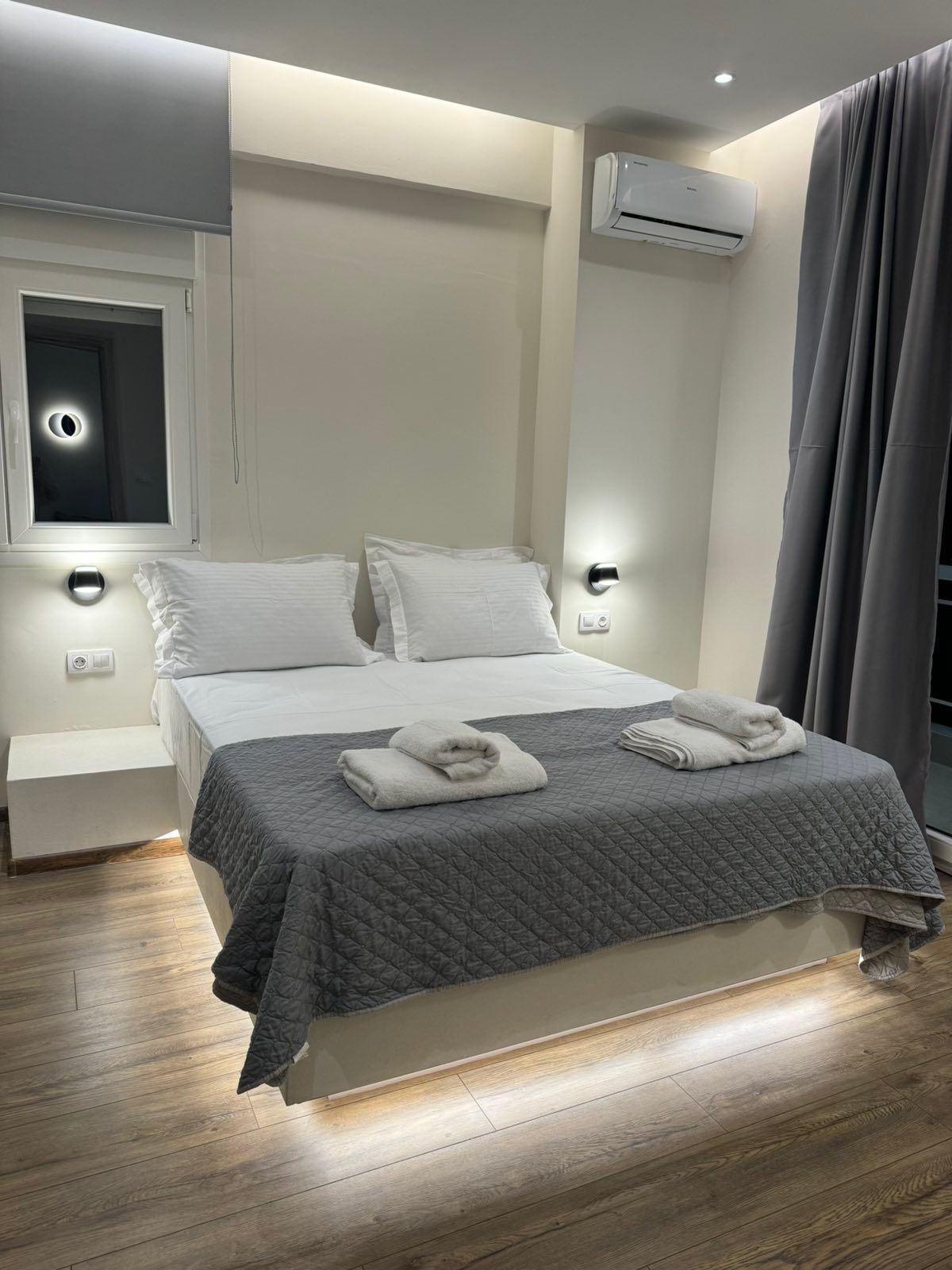 A cozy sleeping area is presented with a spacious bed dressed in neutral tones. Two upholstered pillows are arranged at the head of the bed, with matching towels neatly placed on top. Ambient lighting from wall fixtures and a nearby window adds a touch of brightness.