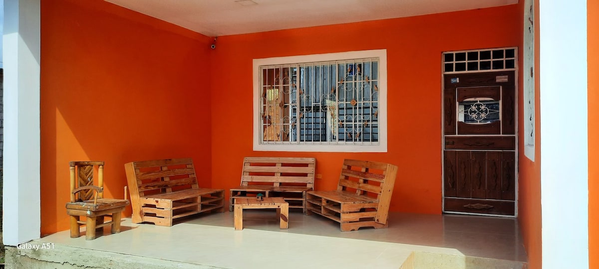 An inviting outdoor seating area features wooden furniture arranged around a central table. The space is framed by vibrant orange walls and a large window with protective bars, allowing natural light to fill the area. The entrance to the home is visible, highlighted by a wooden door.
