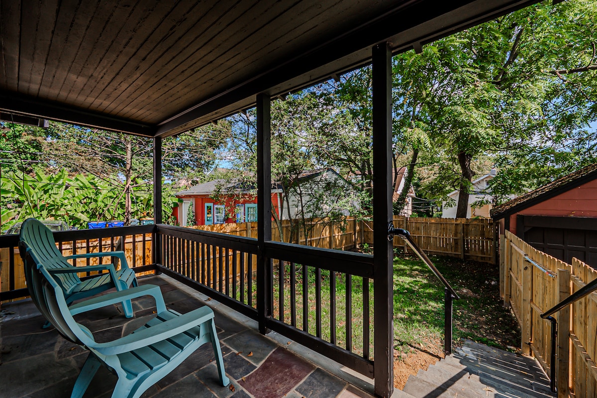 A covered porch features two teal chairs positioned beside a stone floor. A view of the spacious yard is visible through the screened walls, showcasing green grass and trees, along with a fenced area providing privacy.