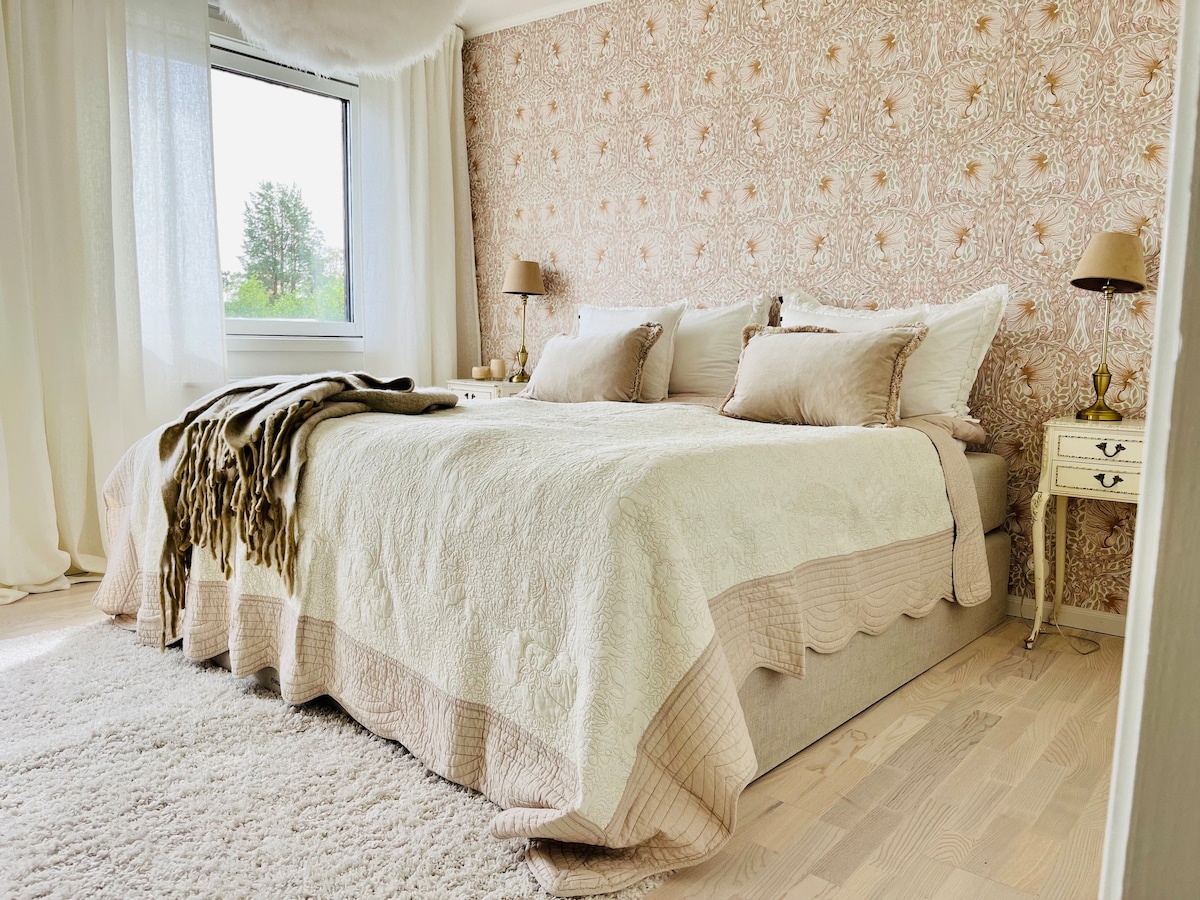 A well-furnished bedroom features a spacious bed with decorative pillows and an elegant quilt. Soft lighting is provided by bedside lamps, while a large window allows natural light to illuminate the room. The textured wallpaper adds a subtle warmth to the serene atmosphere.
