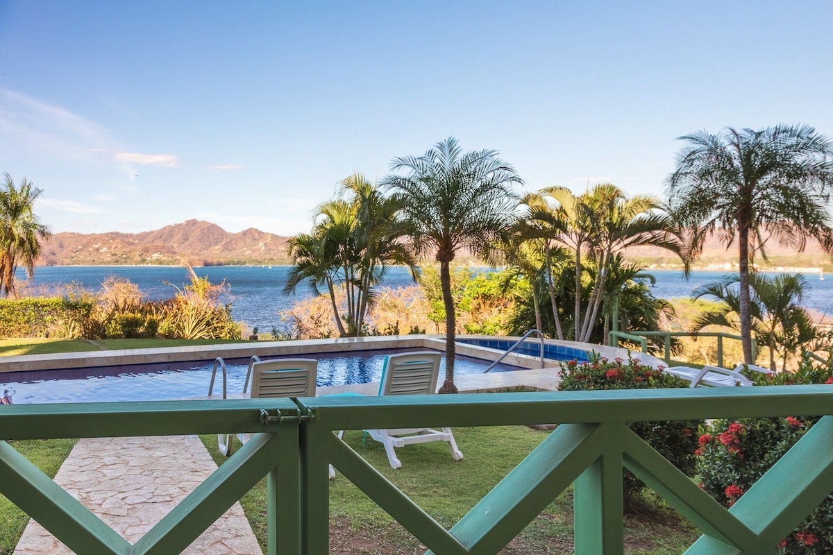 A view showcases a serene landscape featuring a sparkling pool surrounded by lush greenery and palm trees. Beyond the pool, the turquoise water reflects the clear sky and distant mountains, creating a peaceful backdrop for relaxation.