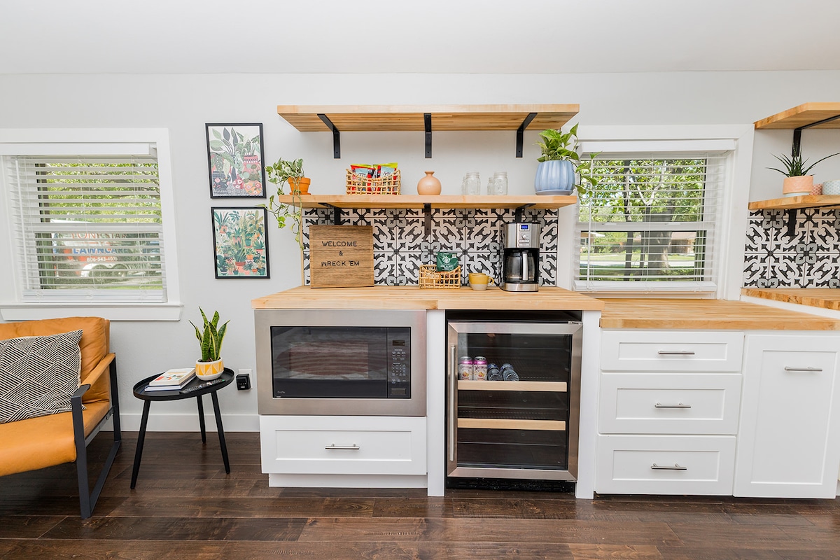 A modern kitchen area features light wood shelves displaying colorful items and plants. The cabinetry includes a microwave and a compact refrigerator. A small table with a chair sits nearby, surrounded by large windows allowing natural light to illuminate the space.