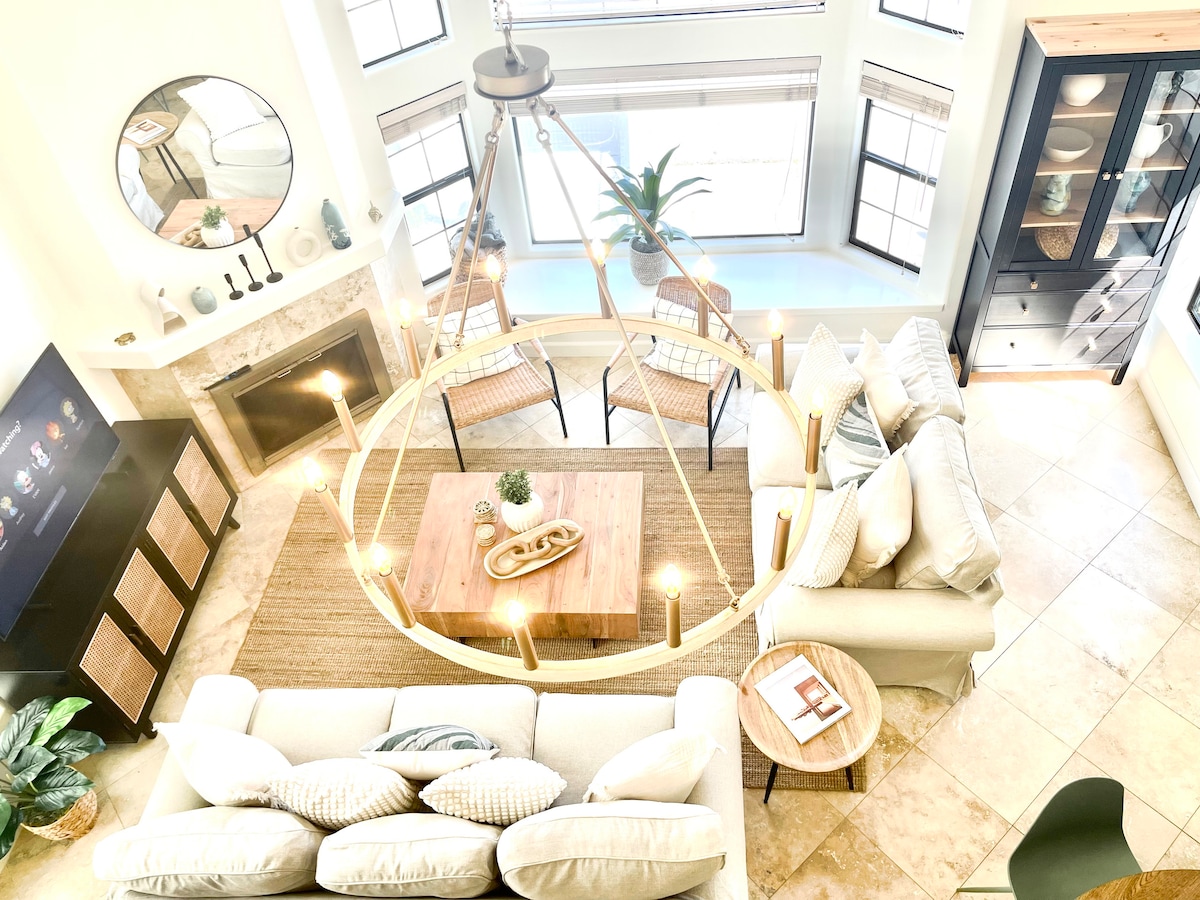 An open living area is highlighted by a round chandelier above a wooden coffee table. A beige sofa and a pair of chairs are arranged around the table. Natural light filters through large windows, illuminating a cozy atmosphere. Decorative plants add a touch of greenery.