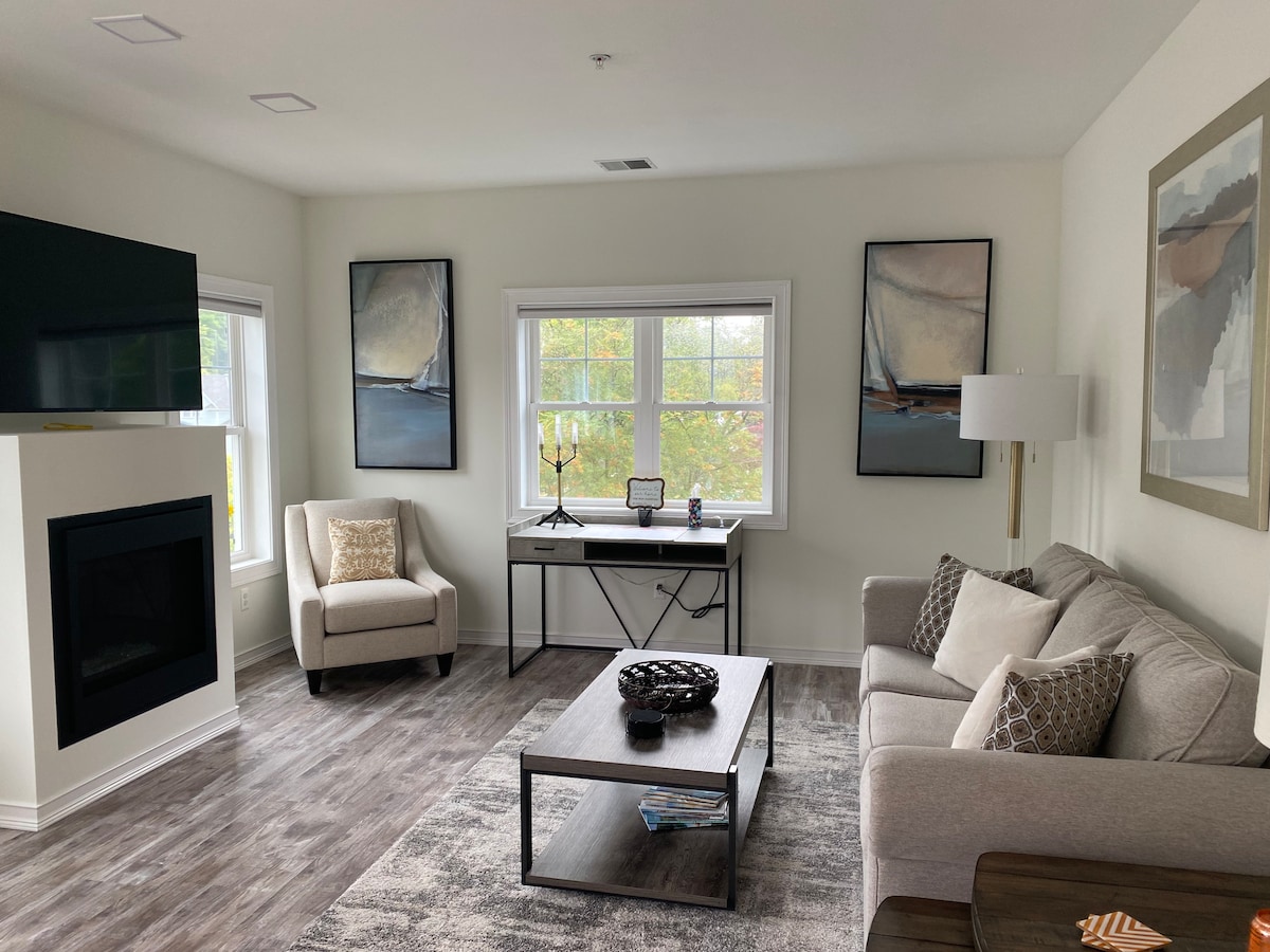 A modern living area features large windows with natural light illuminating the space. A cozy gray sofa and an armchair are complemented by abstract artwork on the walls. A stylish coffee table sits in the center, with a console table positioned against the wall, displaying decorative items.