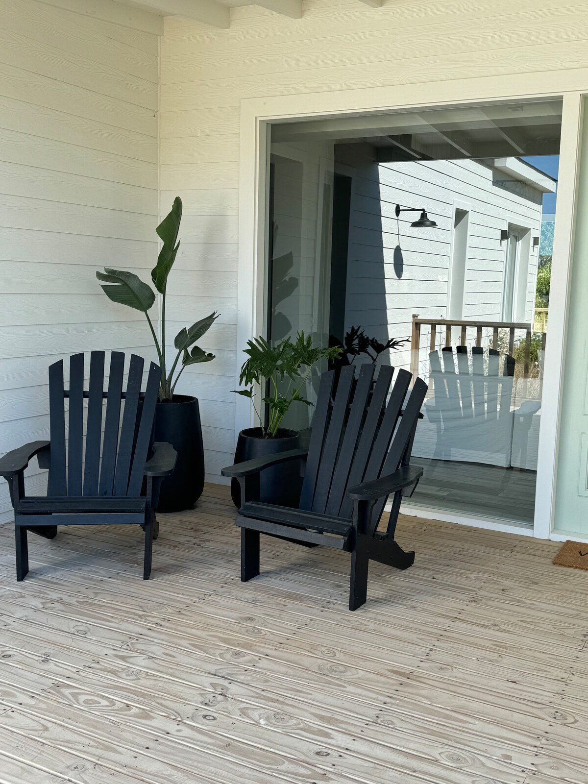 Two black Adirondack chairs are positioned on a wooden deck, accompanied by potted green plants. A large glass door reflects the exterior space, allowing for natural light to enter.