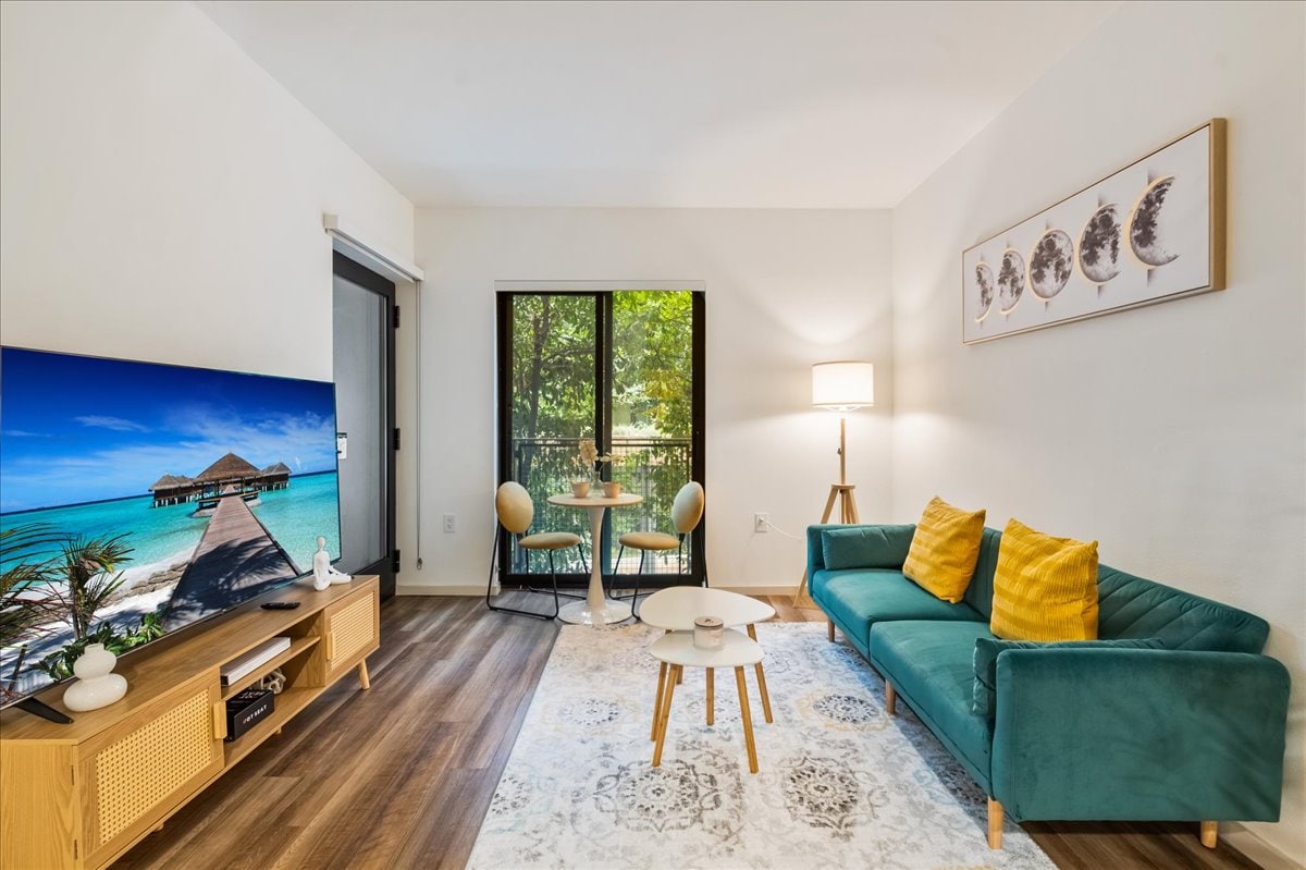 A bright living area features a turquoise couch with yellow accent pillows, paired with a round wooden table and two chairs. A large flat-screen TV displays a scenic beach image. Glass doors provide an outdoor view, while a soft rug adds warmth to the space.