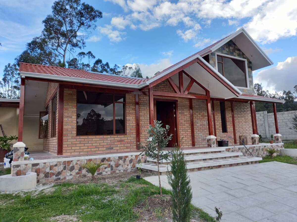 The exterior of the house is showcased, featuring a combination of brick and stone accents. Large windows allow natural light to fill the space, while a covered entryway is supported by wooden beams. Surrounding green areas and a paved pathway add to the inviting atmosphere.