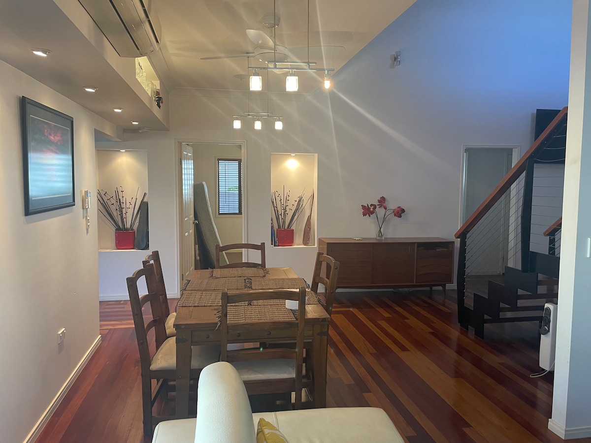An open living and dining area features a wooden table surrounded by chairs, with a variety of decorative elements, including wall art and potted plants. Warm hardwood floors complement the space, with ceiling lights providing illumination. A staircase leads to an upper level.