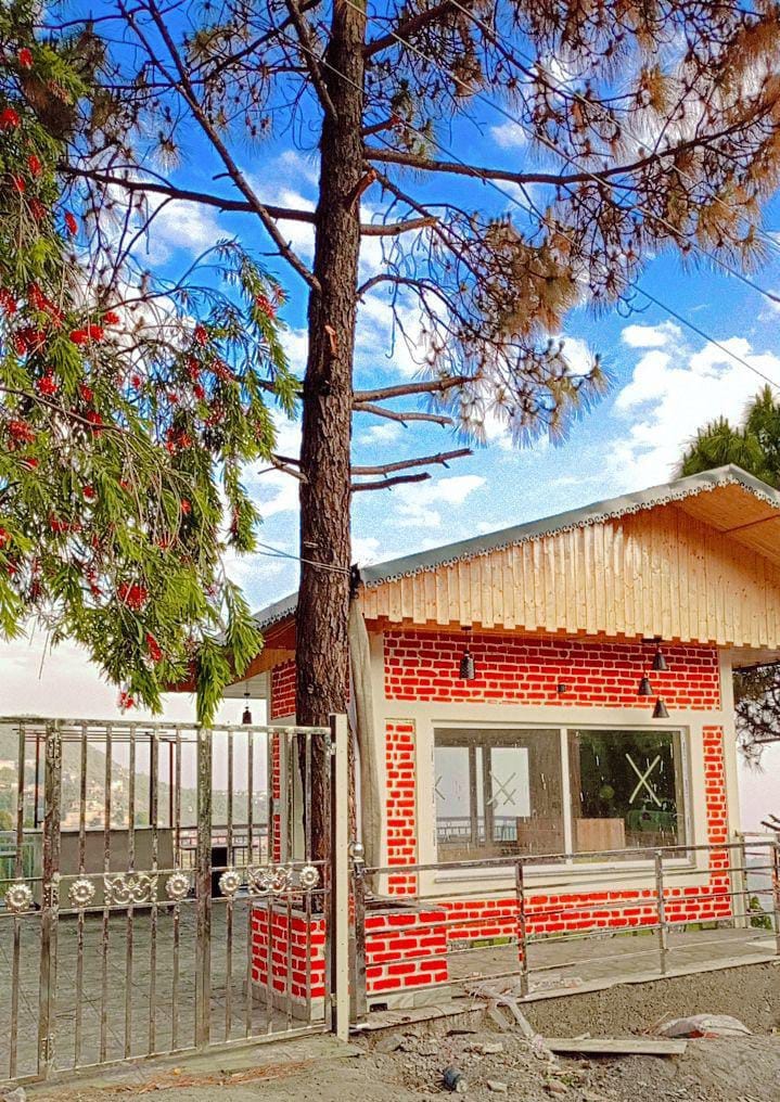 A charming red brick exterior is complemented by a large window, allowing natural light to fill the interior. A tall tree with lush green foliage stands nearby, and the sky above is dotted with soft clouds, contributing to the serene atmosphere.
