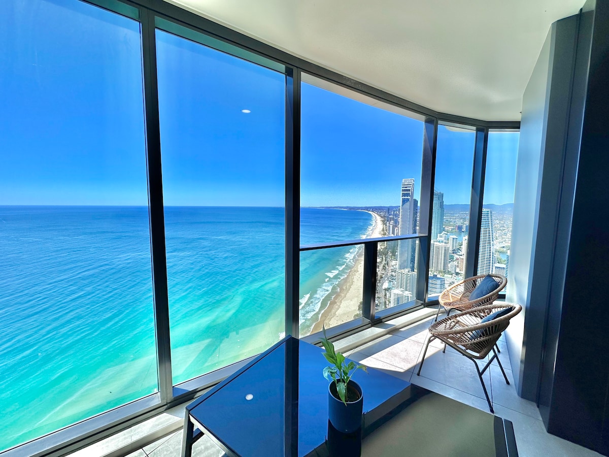A corner unit offers breathtaking 180-degree views of the ocean and city skyline through expansive floor-to-ceiling windows. Two stylish chairs are positioned next to a sleek table, enhancing the modern decor of the living space. Sunlight floods in, reflecting on the vibrant ocean waters.