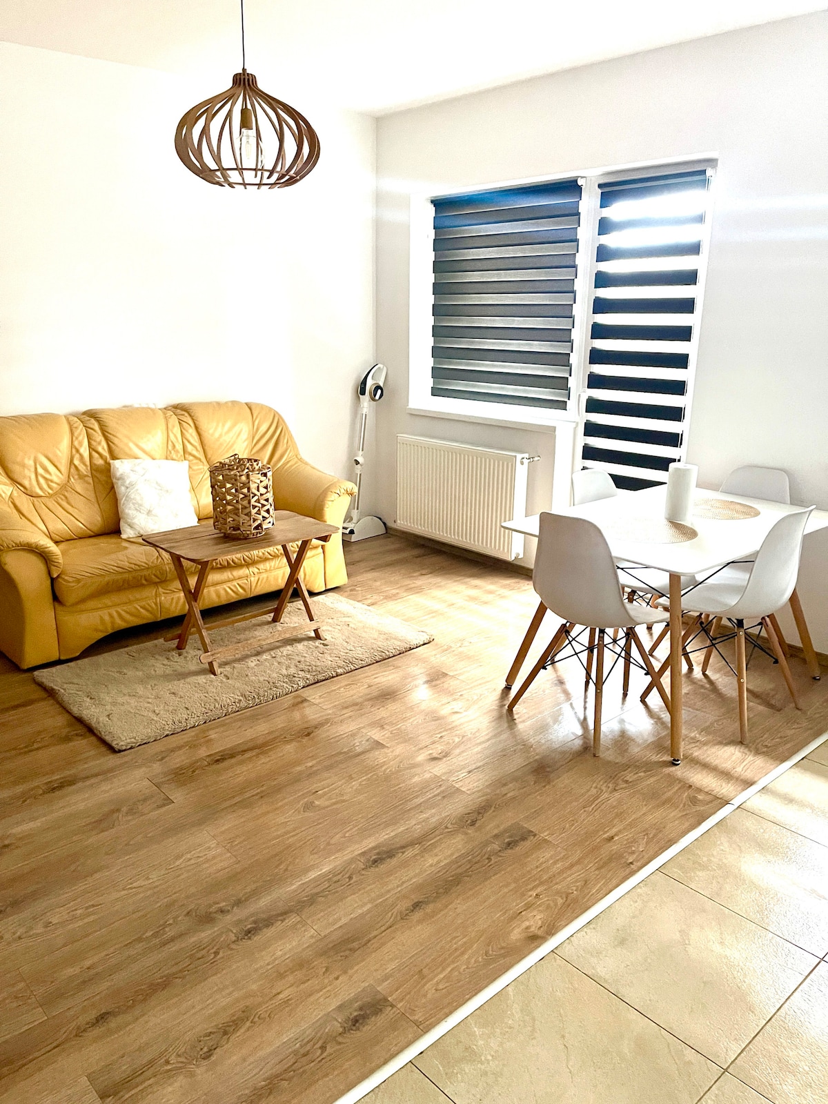 A living area features a yellow sofa paired with a small wooden coffee table. Sunlight filters through the slatted window coverings, illuminating the wooden floor. A dining table with white chairs is positioned nearby, creating a welcoming space for relaxation and meals.