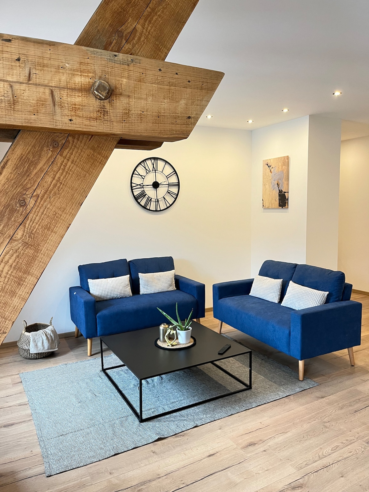 The living area features two blue sofas with decorative pillows, arranged around a black coffee table on a light blue area rug. A wooden beam spans the ceiling, complemented by a wall clock and an abstract piece of artwork on the adjacent wall.