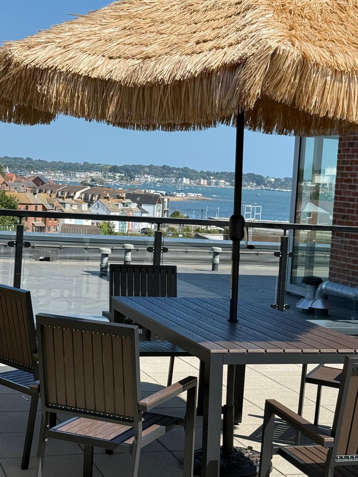 A spacious balcony is depicted with a thatched umbrella providing shade over a dining table surrounded by several chairs. In the background, views of the waterfront and nearby buildings are visible against a clear blue sky.