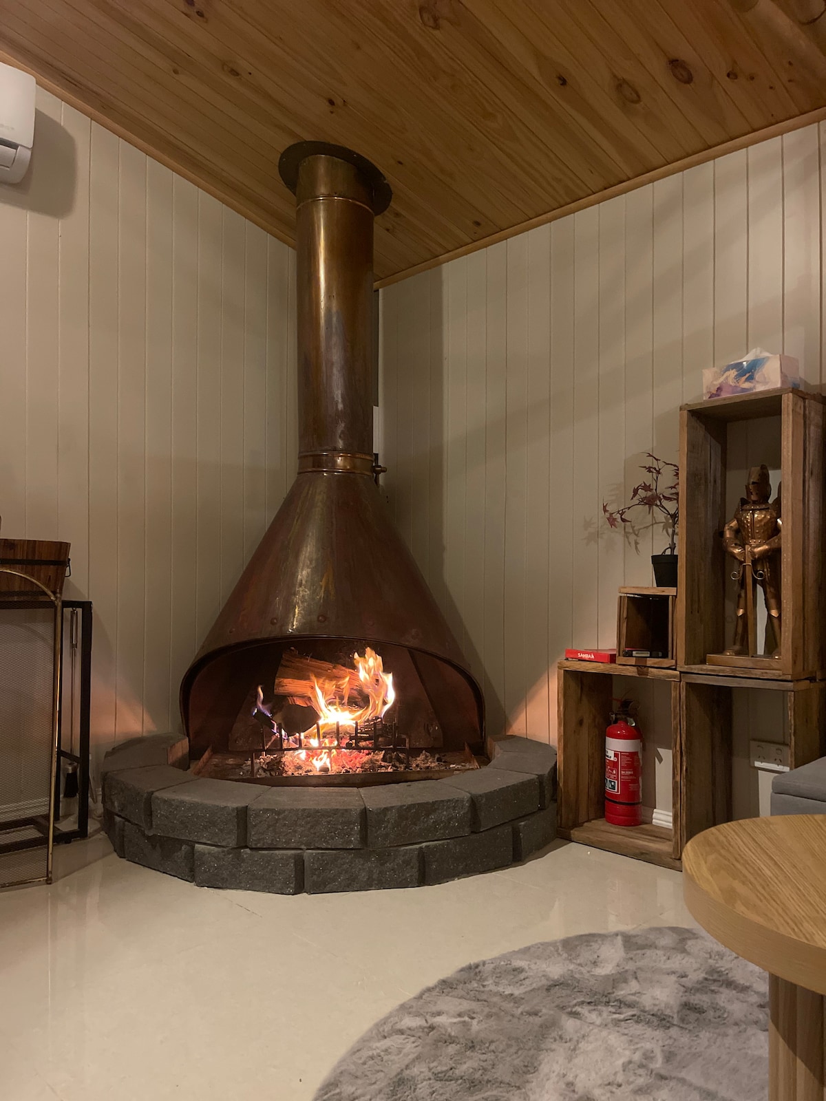 A unique indoor fireplace is featured, set in a circular stone base. Its copper chimney rises to the wooden ceiling, while flames flicker cheerfully. Neatly arranged wooden shelves display decorative items, complemented by a modern side table and a fire extinguisher for safety.