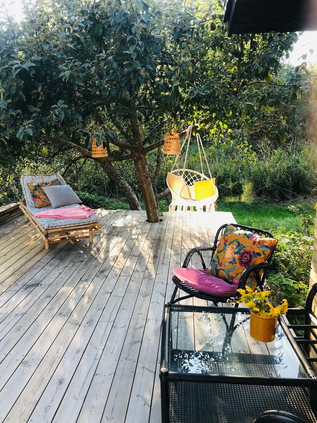 A wooden terrace features a lounge chair with a colorful cushion and a swinging chair suspended from a tree. A glass table with a decorative flower pot is positioned beside a black chair, surrounded by greenery that enhances the serene outdoor setting.