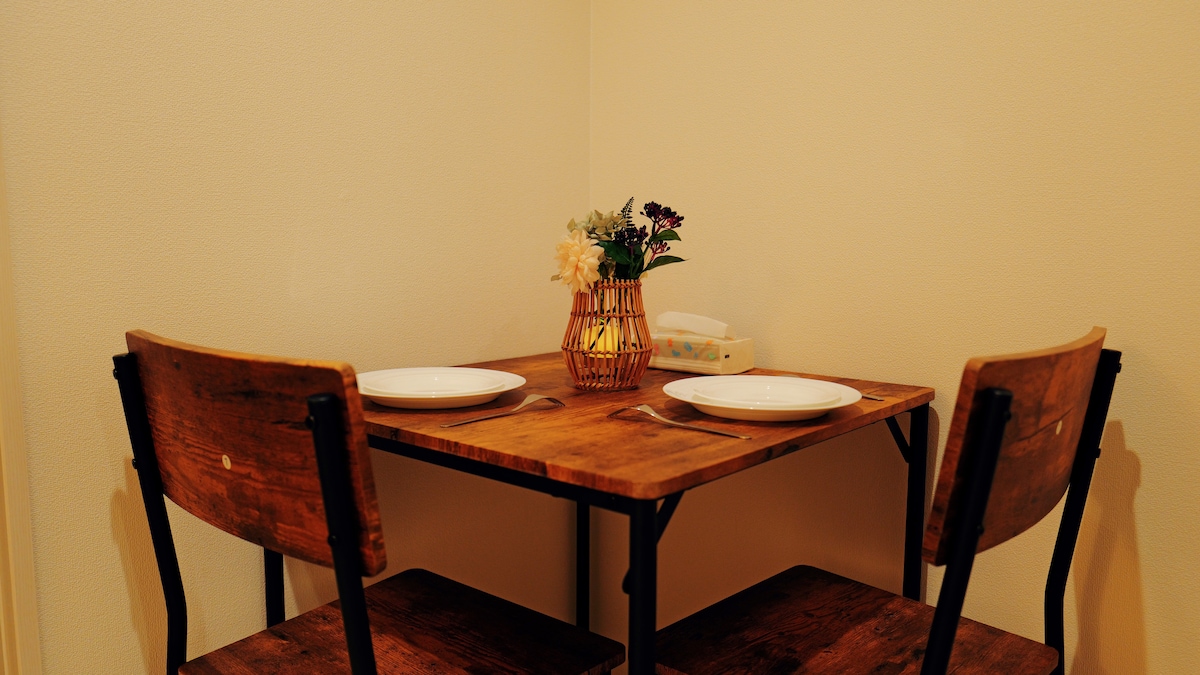 A wooden table is set for two, complemented by two matching chairs. A small vase containing fresh flowers is positioned at the center of the table. The minimalist decor features a neutral wall, enhancing the cozy atmosphere of the dining area.