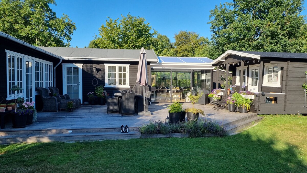 A spacious wooden terrace is surrounded by a green lawn, featuring various potted plants and outdoor seating arrangements. A glassed-in sunroom is visible, providing a light-filled gathering area. Nearby, a charming cabin adds character to the outdoor space, all set against a backdrop of lush trees.