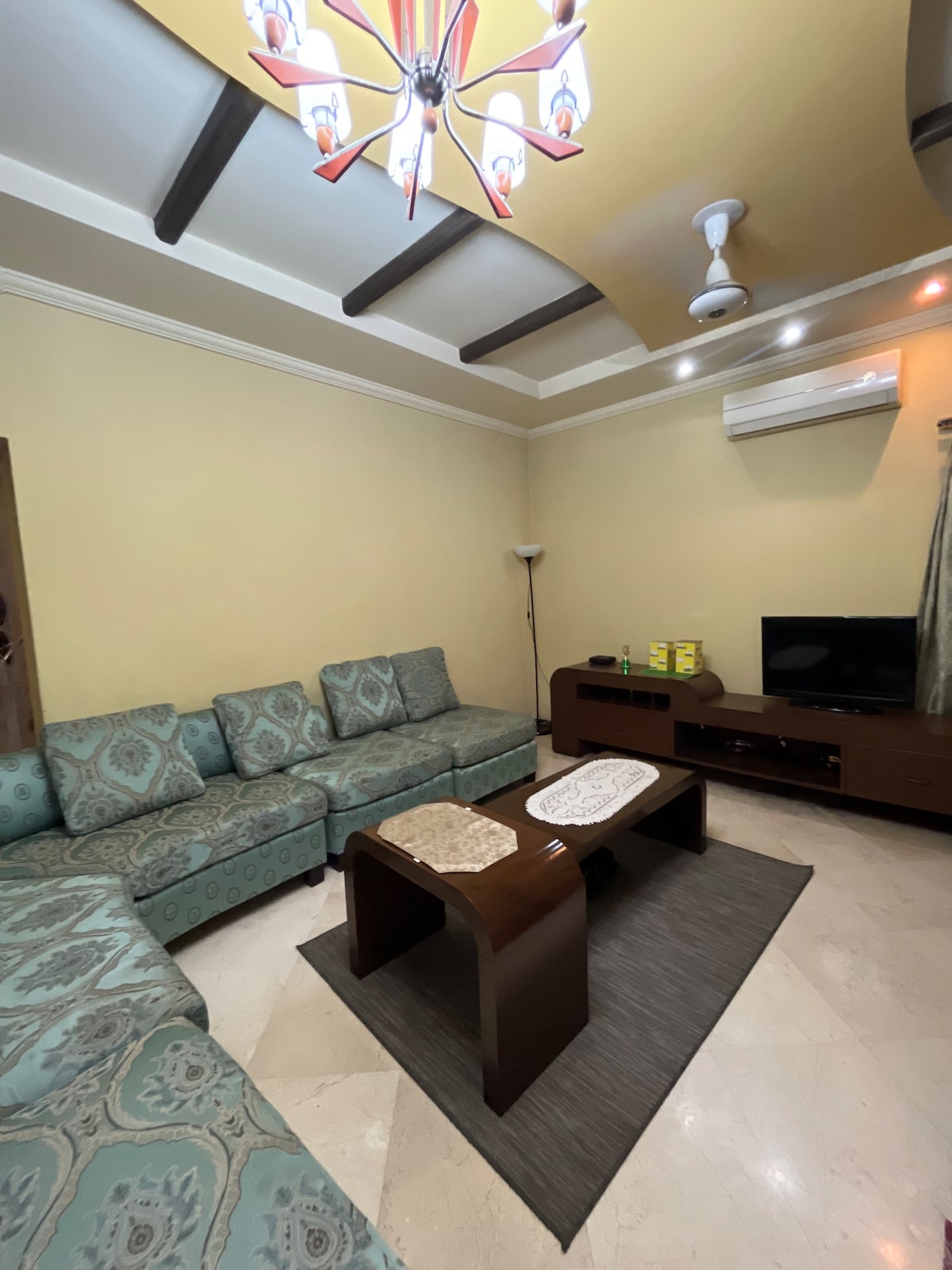A spacious living area features a comfortable sectional sofa arranged around a decorative coffee table. A soft rug lies under the table, while a wall-mounted television is positioned nearby. The room is illuminated by a stylish chandelier and includes a light-colored ceiling with wooden beams.