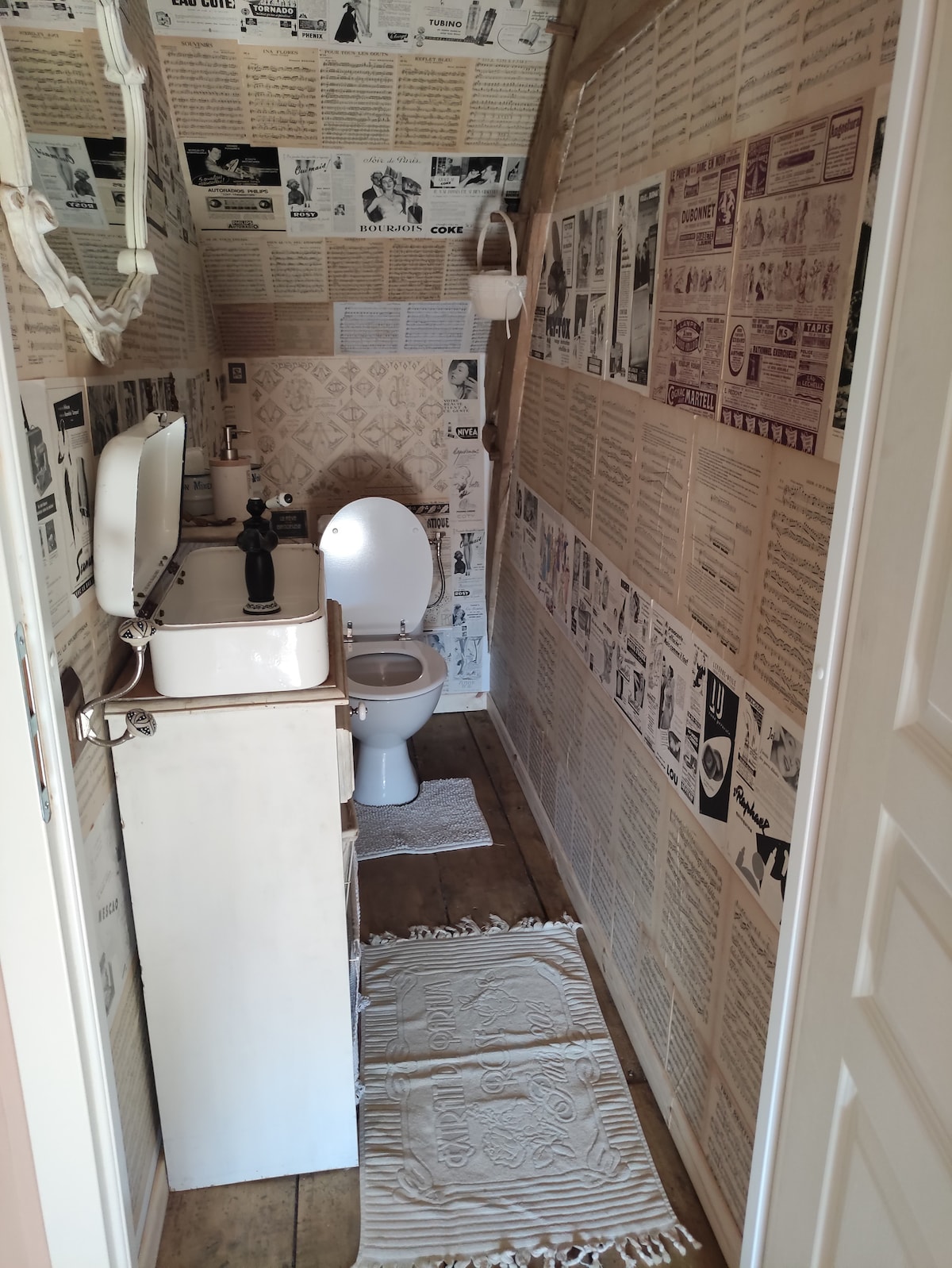 A narrow bathroom is showcased with vintage newspaper wallpaper covering the walls. A white toilet sits adjacent to a wooden sink cabinet, which is complemented by a black faucet. Light-colored bath mats add comfort to the floor, enhancing the space's unique character.
