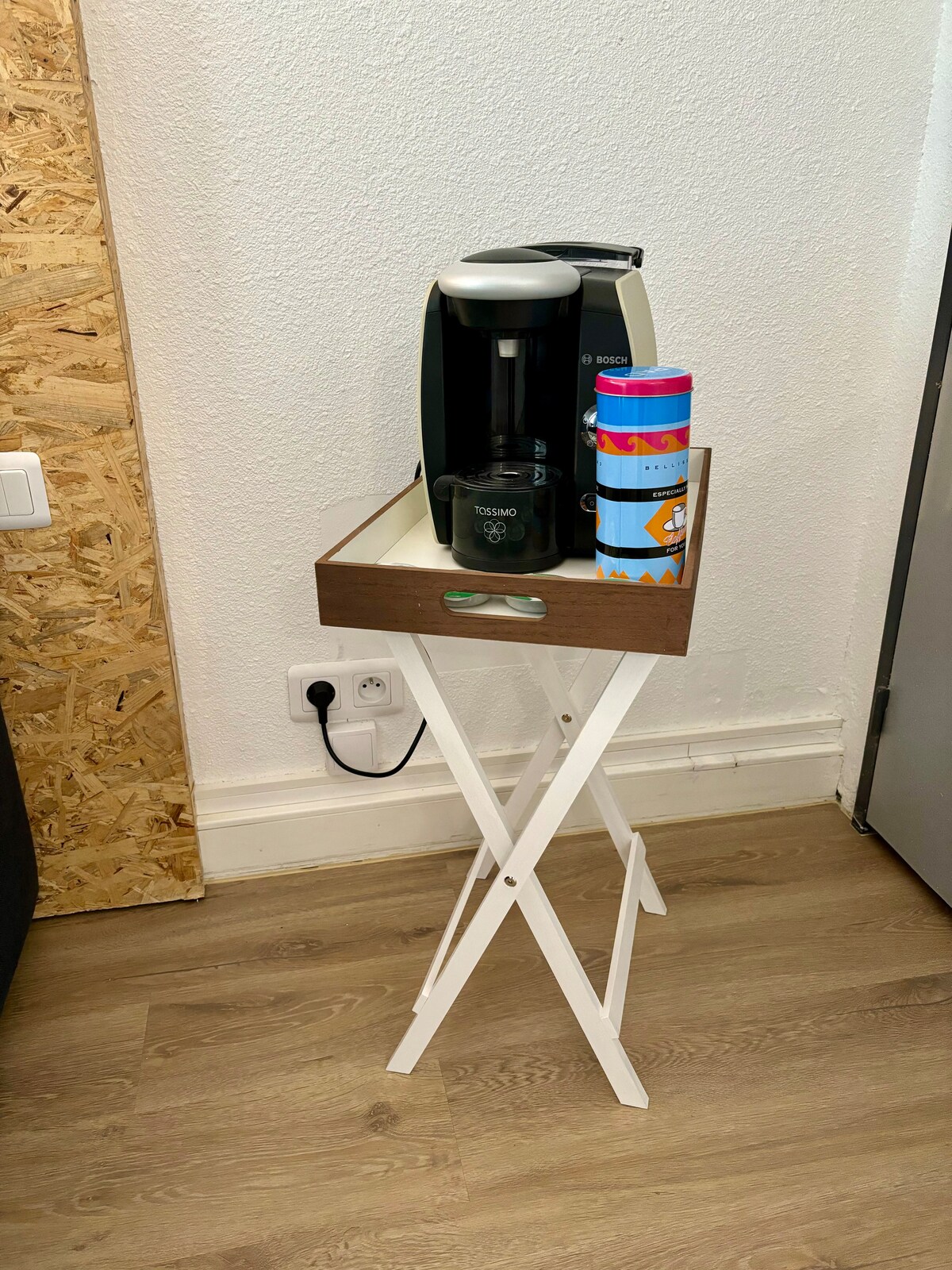 A compact tray table is displayed featuring a coffee machine and colorful travel mugs. The table is set against a light-colored wall, and the wooden surface of the tray contrasts with the white folding legs. Natural light enhances the functional setup.