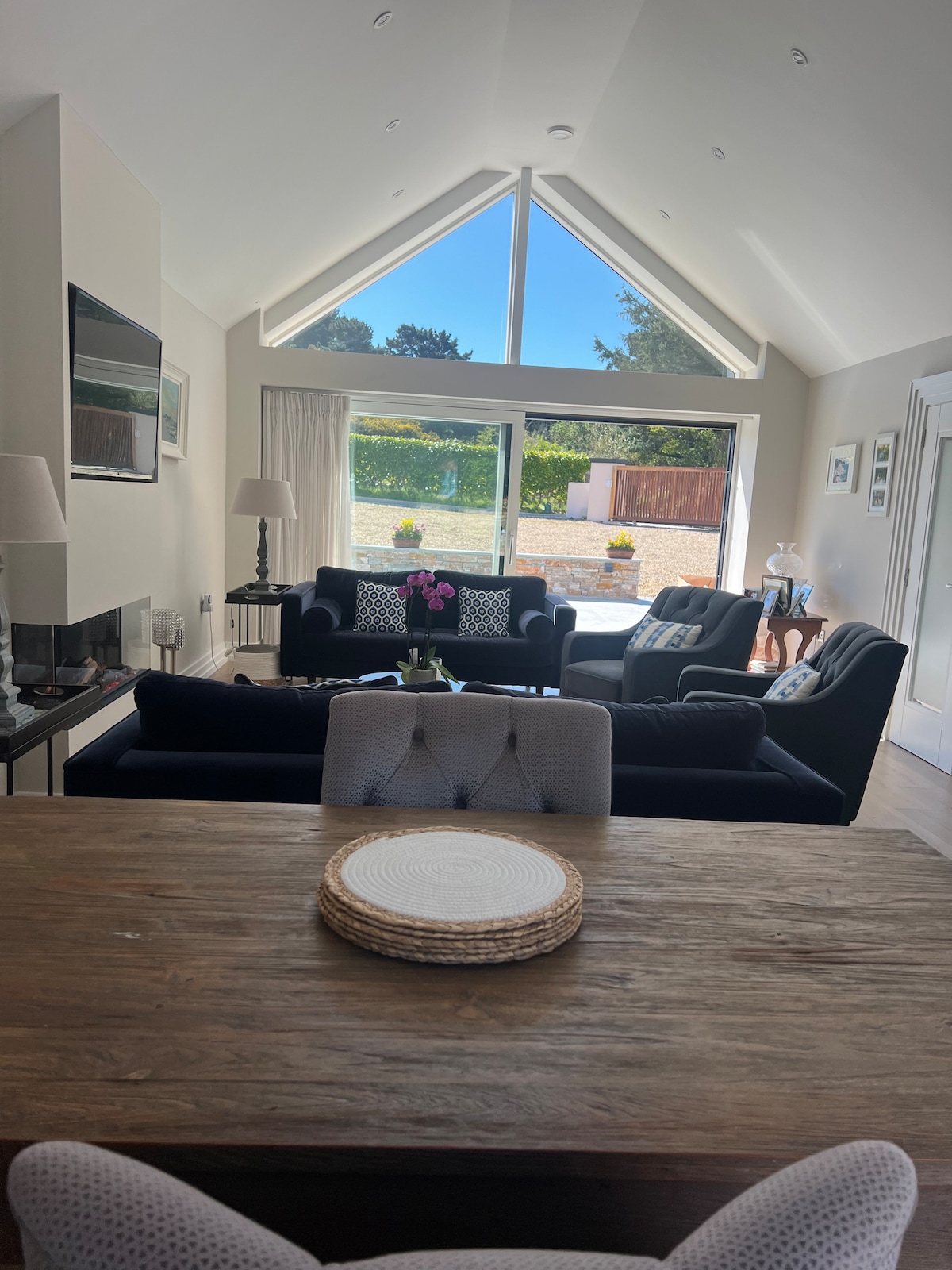 An airy living area features a high, angled ceiling and large windows allowing natural light. Two dark sofas are arranged around a central glass table, while a dining table is visible in the foreground. Colorful plants are positioned on the windowsill, enhancing the space.