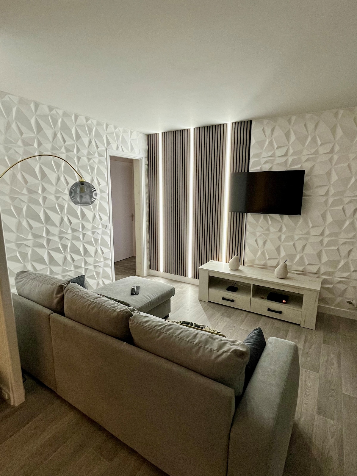 A softly lit living area features a plush sofa facing a wall-mounted television. Modern, textured walls create visual interest, while elegant lighting along the vertical panels adds depth. A small entertainment unit is positioned below the TV, complementing the minimalist decor.