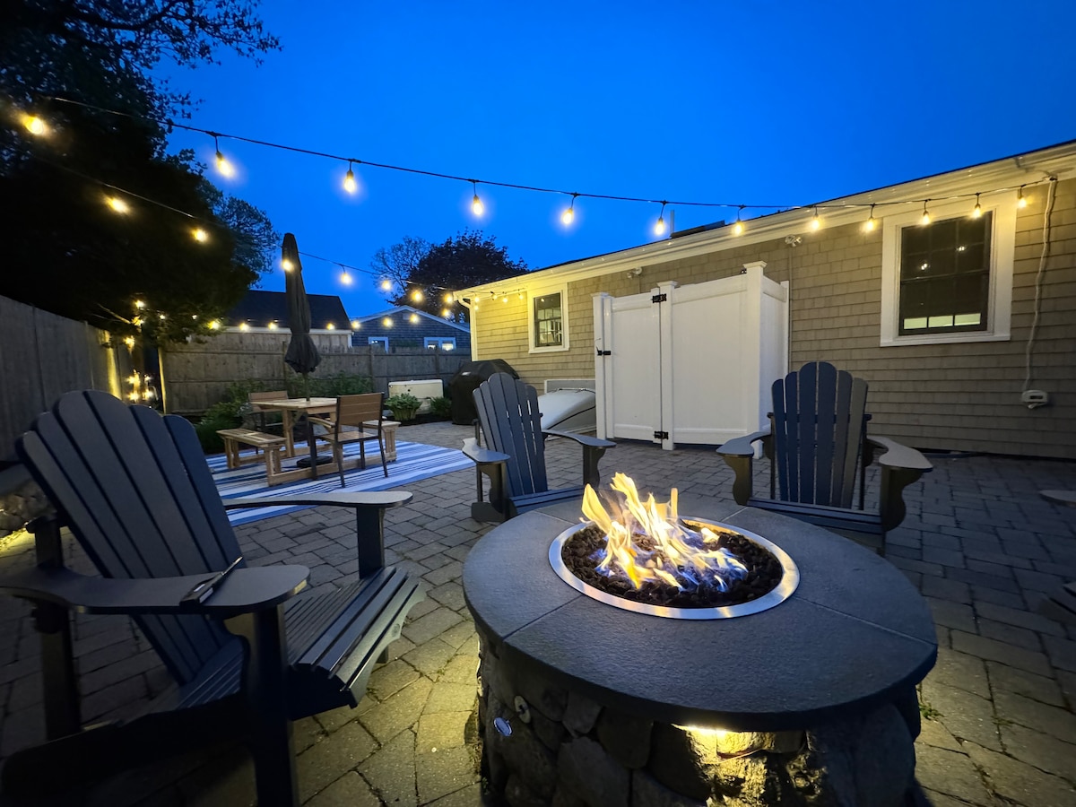 A well-lit outdoor space features four blue Adirondack chairs arranged around a circular stone fire pit. Soft overhead string lights create a cozy ambiance. A dining table with chairs is visible, surrounded by a paved patio, offering an inviting area for relaxation and gatherings.