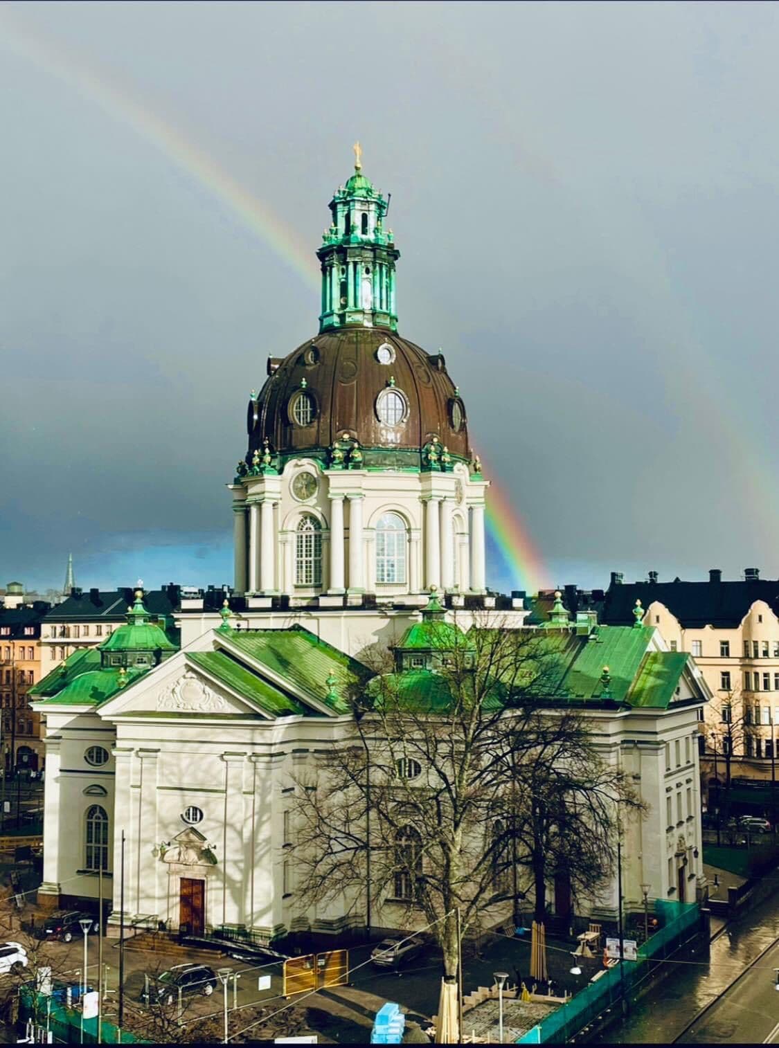 The image captures a prominent architectural structure with a green dome and intricate details. A rainbow arcs across the sky above, enhancing the scene. Surrounding buildings and trees are visible, indicating an urban setting following recent rainfall.