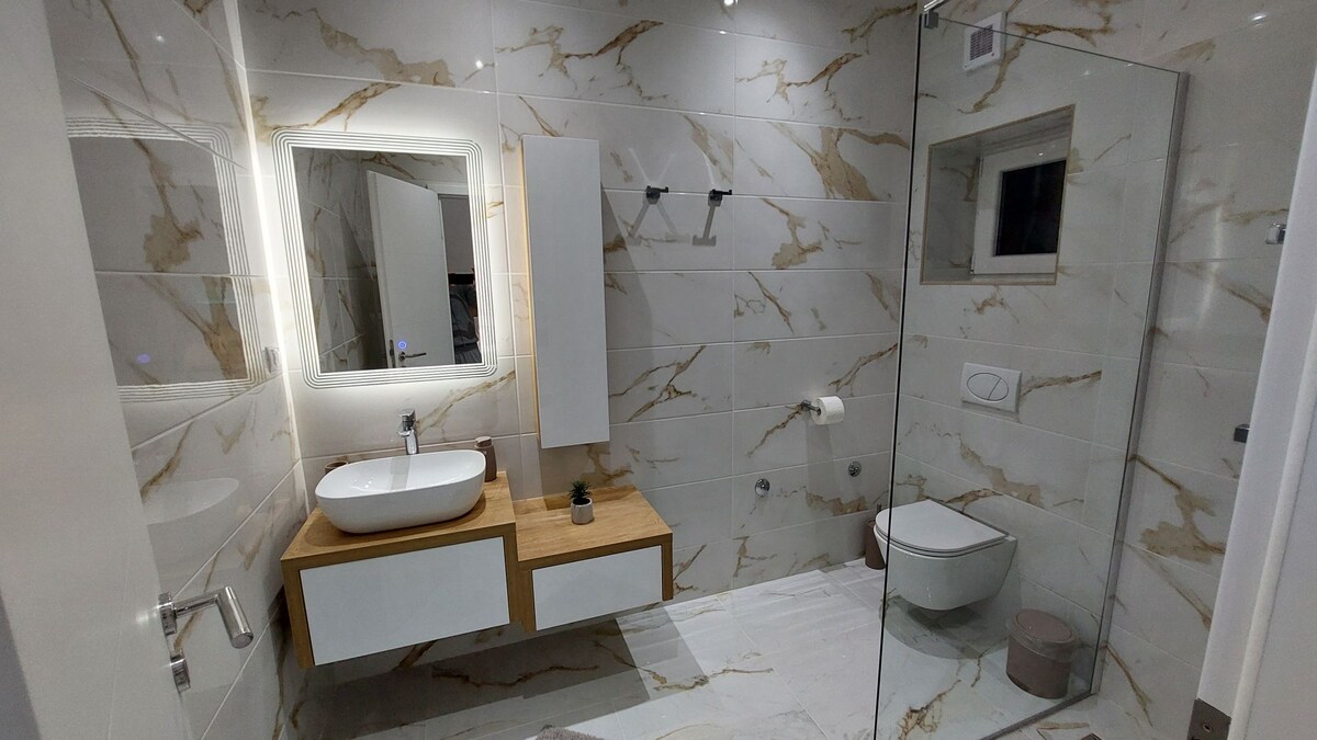 A modern bathroom features elegant marble walls and a stylish lighted mirror. A wooden vanity with a round sink stands against the wall, while a glass shower enclosure is positioned nearby. Minimalistic fixtures and a neutral color palette contribute to a clean and spacious look.