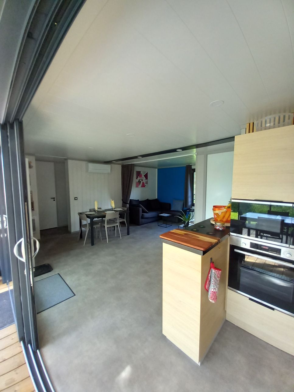 A modern open-plan living area is shown, featuring a dining table with four chairs and a comfortable seating arrangement. A kitchenette is visible with built-in appliances, and large sliding doors provide access to the outdoor space, enhancing natural light within the room.