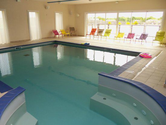 An indoor swimming pool is visible, featuring a smooth, blue-tiled edge and a separate area for relaxation with built-in seating. Colorful lounge chairs are arranged around the pool, and natural light streams in through large windows, enhancing the inviting space.